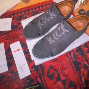  Christian Louboutin - gråa sneakers från Christian Louboutin i mycket bra skick. Skorna har coola nitar på tån och den ikoniska röda sulan. Perfekta för att ge din outfit en edgy touch! De är både stiliga och bekväma, med snörning för perfekt passform.