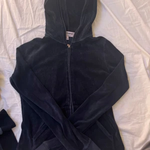 Svart hoodie från Juicy Couture - Mysig svart hoodie från Juicy Couture i mjukt material. Den har en dragkedja framtill och en praktisk huva. Perfekt för en avslappnad stil.