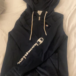 Mörkblå hoodie från Champion - Snygg svart hoodie från Champion med dragkedja och vit logga på ärmen. Ganska tunt material perfekt under en jacka, eller ett linne under hoodien. Strlk XS men passar S. Säljs inte längre men köpt för ungefär 550kr.