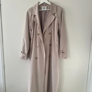 Beige trenchcoat från JDY - Snygg beige trenchcoat från JDY med klassisk dubbelknäppning och knytskärp i midjan. Jackan har långa ärmar och stilrena knappar på ärmsluten. Perfekt för en stilren look. Har inte hunnit använda den så säljer den nu. Nypris 799