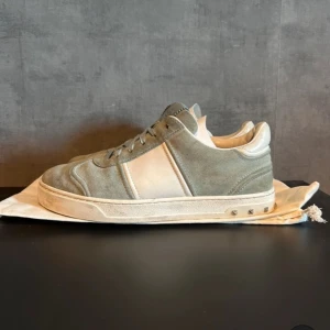 Grå sneakers från Valentino - Säljer ett par snygga grå sneakers från Valentino i mocka och skinn. De har en stilren design med snörning och coola nitar på hälen. Perfekta för både vardag och fest! Skorna är i bra skick och kommer med originalpåse. Tar också emot byte. Skorna är storlek 38,5 men passar 39,39,5 och om du löser snören kanske till och med strlk 40