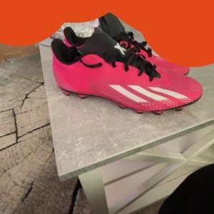 Rosa fotbollsskor från Adidas - Säljer ett par snygga rosa fotbollsskor från Adidas i mycket bra skick. Skorna har en cool design med vita detaljer och svarta snören. Perfekta för fotbollsträning eller match. De har en slitstark sula med dobbar för bra grepp på planen. Passar dig som vill sticka ut på planen!
