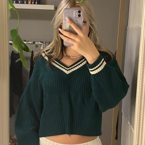 Mörkgrön stickad tröja med V-ringning - Säljer en superfin mörkgrön stickad tröja med V-ringning och vita ränder vid halsen och ärmsluten. Tröjan är croppad och har en loose passform, perfekt för höst och vår. Den är i mycket bra skick och passar perfekt till både jeans och kjol. Perfekt för en mysig dag ute eller hemma!