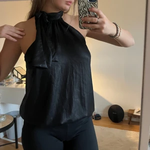 Svart halterneck-top - Snygg svart halterneck-top i glansigt material med knytning vid halsen. Perfekt för en utekväll eller fest. Toppen har en lös passform och är ärmlös, står inte storlek men skulle säga S/M💖