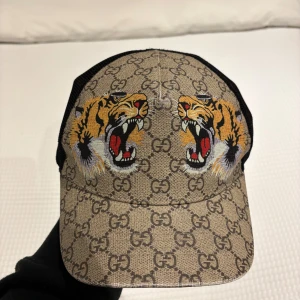 Mönstrad keps med tigerbroderi - Säljer en snygg keps från Gucci med ett coolt tigerbroderi på framsidan. Kepsen har ett klassiskt GG-mönster i beige och svart, och är tillverkad i bomull med nätmaterial på baksidan för extra ventilation. Perfekt för att ge din outfit en lyxig touch!