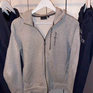 Grå hoodie från Sail Racing - Säljer en grå hoodie från Sail Racing i storlek 160. Den har en dragkedja framtill och en liten ficka på bröstet men även en mysig huva. Perfekt för både höst och vår. Materialet är en blandning av bomull och polyester, vilket gör den både bekväm och hållbar. Passar perfekt för en avslappnad stil! Skriv för funderingar. PRIS EJ HUGGET I STEN!