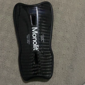 Svart Monolit skydd - Säljer ett svart Monolit skydd i kolfiber. Det är lätt och har en ergonomisk form för bästa komfort. Perfekt för träning eller match. Skyddet är i mycket bra skick och redo för användning.