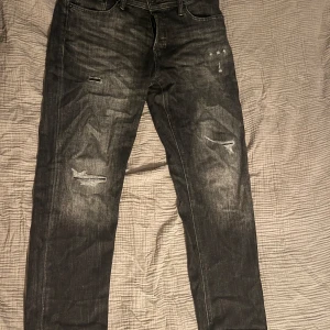 Svarta jeans från Jack & Jones - Säljer ett par svarta jeans från Jack & Jones i modellen Tapered/Mike. De har en cool slitningseffekt och är i bra skick. Perfekta för en avslappnad stil med en touch av edge. Passar både till vardags och fest!