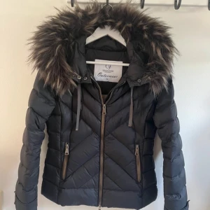 Svart dunjacka - Snygg svart dunjacka från Chiara Forthi med fluffig fuskpäls på huvan. Jackan har en quiltad design och dragkedja framtill samt två praktiska fickor med dragkedjor. Perfekt för kyliga höst- och vinterdagar. Håll dig varm och stilren! 🖤