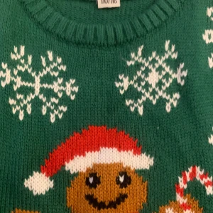 Grön jultröja med pepparkaksgubbe - Supermysig grön jultröja från North med en söt pepparkaksgubbe i tomteluva och texten 'Happy Holidays'. Tröjan har snöflingor som mönster och är perfekt för julfirandet. Den är stickad och har långa ärmar, vilket gör den både varm och bekväm. Perfekt för att sprida julstämning! 🎄