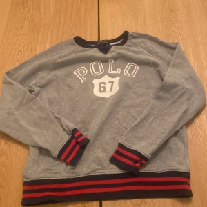 Grå tröja från Ralph Lauren - Säljer en snygg grå tröja från Ralph Lauren med texten 'POLO 67' på framsidan. Tröjan har långa ärmar och ribbade muddar i rött och blått vid ärmslut och nederkant. Perfekt för en avslappnad stil! Har ett litet hål i ryggen men det syns knappt 