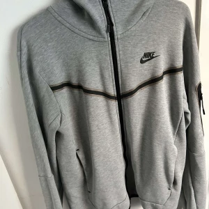 Grå hoodie från Nike - Säljer en grå hoodie från Nike i bra skick. Den har en dragkedja framtill och en svart randdetalj över bröstet. Perfekt för höst och vår, med långa ärmar och en bekväm passform. Huvan ger extra mysfaktor. Passar både till träning och vardag.