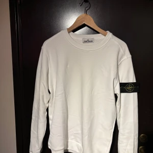 Vit långärmad tröja från Stone Island - Säljer en stilren vit långärmad tröja från Stone Island. Den har en klassisk design med rund hals och det ikoniska märket på ärmen. Perfekt för både vardag och festliga tillfällen. Tröjan är i mycket bra skick och redo för en ny ägare!