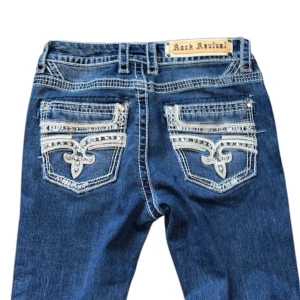 Rock Revival jeans - Snygga jeans från Rock Revival i mörkblå denim med unika broderade detaljer på bakfickorna. De har en bootcut-stil och är perfekt för en avslappnad men ändå stilren look. Jeansen har en låg midja och är i bra skick. Perfekta för både vardag och fest! Storlek S, midja är 35cm och innerben är 79cm