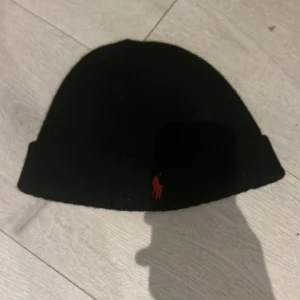 Svart mössa från Polo Ralph Lauren - Snygg svart mössa från Polo Ralph Lauren med det klassiska röda broderade logotypen framtill. Perfekt för att hålla värmen under kalla dagar. Tillverkad i mjukt och bekvämt material. Passar till alla outfits och är ett måste för vintern!