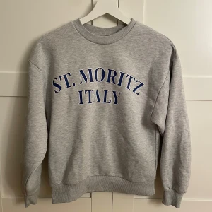 Grå sweatshirt från Gina Tricot - Säljer en snygg grå sweatshirt från Gina Tricot i storlek XXS. Den har texten 'ST. MORITZ ITALY' i blått på framsidan. Perfekt för en avslappnad stil och passar till alla årstider. Tröjan är i bra skick men är lite nopprig som man kan se på en av bilderna men inget man tänker på❤️Skriv om ni har några frågor❤️