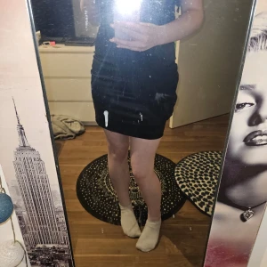 Svart axelbandslös klänning - Snygg svart axelbandslös klänning i satin med omlott-detaljer. Perfekt för festliga tillfällen. Klänningen har en elegant drapering framtill och en dragkedja bak för enkel påklädning.