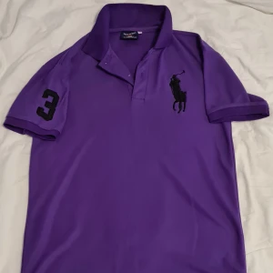 Lila pikétröja från Polo Sport - Säljer en snygg lila pikétröja från Polo Sport. Den har en klassisk krage och korta ärmar med en svart siffra '3' på ena ärmen. Perfekt för en sportig och stilren look. Passar både till vardags och lite finare tillfällen. 🏇