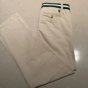 Beige chinos från Polo Ralph Lauren - Säljer ett par snygga beige chinos från Polo Ralph Lauren. Storleken är 12 år och sitter som slim. De har en klassisk design med dragkedja och knapp framtill. Byxorna kommer med ett bälte som jag fick när jag köpte byxorna. Nypris är runt 800 kr. Varan är inprincip helt ny och har bara använts runt fem gånger.
