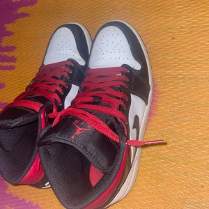 Nike Air Jordans med röd och svart design - Säljer ett par snygga Nike Air Jordans i svart, vitt och rött. Skorna har en klassisk design med röd snörning och den ikoniska Nike-loggan på sidan. Perfekta för både vardag och fest. De är i bra skick och redo för nya äventyr!