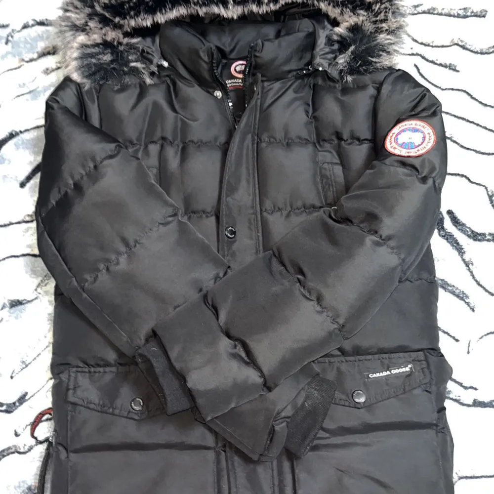 Säljer en svart dunjacka från Canada Goose i bra skick. Jackan har en fluffig pälsfodrad huva och är perfekt för kalla vinterdagar. Den har en dragkedja och knappar framtill för extra skydd mot kylan. Perfekt för vintern! ❄️ Det är en knapp som har ramlat av . Jackor.