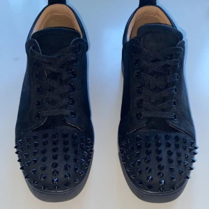 Christian Louboutin   - Säljer ett par svarta sneakers från Christian Louboutin i mocka med coola nitar på tån. De har en klassisk röd sula och snörning. Perfekta för att ge din outfit en edgy touch! Använda men i bra skick. Höger skon har tappat 3 skruvar och vänster skon 1 skruv, men det är absolut inget man märker av. Därför jag säljer billigt!! Först till kvarn!