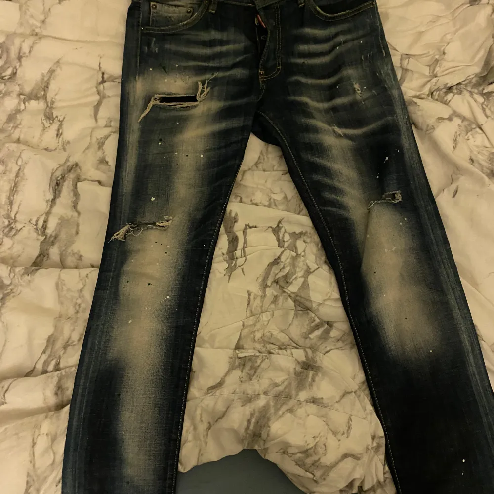 Säljer ett par snygga blå jeans från Dsquared2 i bra skick. De har en cool slitning och färgstänk som ger en unik look. Perfekta för både vardag och fest! Passformen är normal och de är tillverkade i bomull för en skön känsla.. Jeans & Byxor.