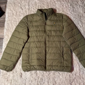 Olivgrön quiltad jacka från Lyle & Scott - Säljer en olivgrön quiltad jacka från Lyle & Scott i mycket bra skick. Jackan har en dragkedja framtill och ett broderat emblem på bröstet. Perfekt för höst och vår med sin stilrena design och bekväma passform. Passar ungdomar i åldern 15-16 år.