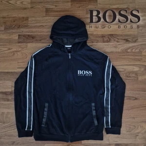 Svart hoodie från Hugo Boss - Säljer en snygg oanvänd hoodie från Hugo Boss i mycket bra skick. Den har en dragkedja framtill och en huva för extra komfort. Perfekt för höst och vår, med vita detaljer längs ärmarna som ger en sportig look. Det är en eprfekt julklapp åt dig själv eller nära och kära🎁 Passa på!