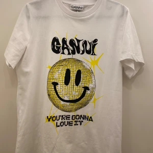 Vit t-shirt från Ganni  - Säljer en supercool vit t-shirt från Ganni i storlek XS. Den har ett stort gult smileytryck med texten 'YOU'RE GONNA LOVE IT'. Endast testad! 