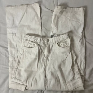 Vita cargopants - Säljer ett par snygga vita cargopants i mycket bra skick. De har en dragkedja och knappar framtill samt praktiska fickor på sidorna. Perfekta för en avslappnad stil och passar till de flesta tillfällen. Storleken är inte angiven, men de ser ut att vara i en normal passform. Dock lite smutsigt där nere men de går bort vid tvätt.