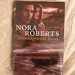 Främlingen vid havet - Spännande bok av Nora Roberts, 'Främlingen vid havet', utgiven av Albert Bonniers Förlag. Omslaget visar en dramatisk kustscen med en man och kvinna i förgrunden, vilket ger en mystisk känsla. Perfekt för dig som gillar romantik och spänning!