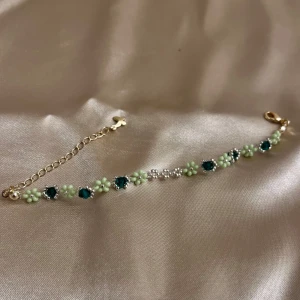 Blommigt armband med gröna och blå detaljer - Ett superfint armband med små blommor i ljusgrönt och gnistrande blå stenar. Armbandet har en justerbar kedja i guldton och 14k guld pläterad som gör det enkelt att anpassa storleken. Perfekt för att ge en färgklick till din outfit! 