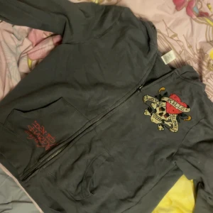 Grå hoodie från Ed Hardy - Säljer en grå hoodie från Ed Hardy med coolt tryck av en dödskalle och texten 'Love Kills Slowly'. Den har en dragkedja framtill och en liten reva på ena sidan. Perfekt för en avslappnad stil med attityd! Har dock ett litet hål vid fickan! och små fläckar runt om kring på tröjan därav pris