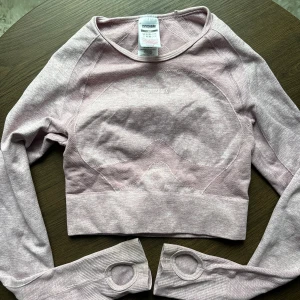 Ljusrosa croppad tröja från Gymshark - Säljer en superfin ljusrosa croppad tröja från Gymshark. Perfekt för träning med sin tighta passform och långa ärmar. Den har en snygg öppen ryggdetalj och tumhål vid ärmsluten. Materialet är mjukt och stretchigt, vilket gör den bekväm att bära. Passar perfekt för gymmet eller en aktiv dag!