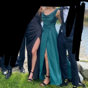 Grön balklänning med slits - Snygg grön långklänning med en elegant slits framtill. Klänningen har en vacker draperad halslinje och är ärmlös, perfekt för festliga tillfällen. Materialet ser ut att vara av satin vilket ger en fin glans. Perfekt för bal eller andra speciella tillfällen!
