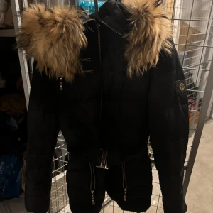 Svart dunjacka med pälsdetaljer - Säljer en snygg svart dunjacka med fluffig päls (äkta)  på huvan. Jackan har dragkedja ska dock sägas att ”flärpen” inte själva draggrejen men flärpen som är på är borta. Mycket bra vinterjacka för någon som fryser som mig! Förutom ”flärpen” är jackan i fint skick, säls för jag inte använder den längre 