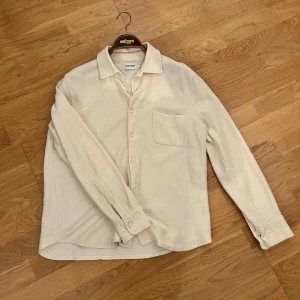 Vit overskirt från East West - Säljer en riktigt fet overskirt från East West i mycket bra skick. Skjortan har långa ärmar och en klassisk krage med knappar framtill. Perfekt för både vardag och festliga tillfällen. Materialet känns mjukt och bekvämt, vilket gör den idealisk för vår och höst. Passar bra till jeans eller chinos för en avslappnad look. Nästan helt oanvänd på grund av att den var för liten.