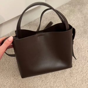 Brun axelväska i skinn - Säljer denna super fina väskan från ARKET den större modellen i en fin mörkbrun färg använd ett fåtal gånger därav mycket bra skick💕