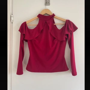 Röd topp med volangdetaljer från Nelly - Snygg röd topp från Nelly i storlek M. Den har långa ärmar och en unik design med volangdetaljer och öppna axlar. Perfekt för festliga tillfällen eller en utekväll.