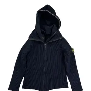 Säljer en snygg svart ribbad hoodie från Stone Island. Den har en dragkedja framtill och en skön huva. Perfekt för höst och vinter, med den klassiska loggan på ärmen. Passar både till vardags och lite mer avslappnade tillfällen. Grymt bra skick! Man kan fälla in luvan i ryggen om önskas. Storlek XS