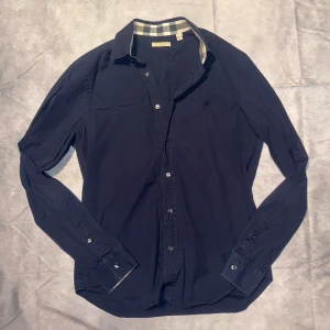 Burberry Skjorta - Säljer en snygg mörkblå skjorta från Burberry i bomull. Den har långa ärmar och klassiska knappar framtill. Kragen har det ikoniska Burberry-rutiga mönstret på insidan. Perfekt för både vardag och fest! Storlek M.