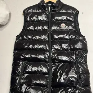 Säljer en snygg svart glansig dunväst från Moncler. Västen har en dragkedja framtill och två praktiska fickor med dragkedjor. Perfekt för kyliga dagar och ger en stilren look. Passar både till vardags och festliga tillfällen. Har ett litet hål på höger fickan 