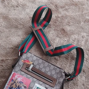 Axelväska med mönster och broderad detalj - Säljer en snygg axelväska med ett klassiskt mönster och en cool broderad detalj på framsidan. Väskan har en justerbar axelrem i grönt och rött, samt en praktisk dragkedja framtill. Perfekt för vardagsbruk eller en dag på stan!