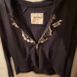 Svart hoodie från Abercrombie - Säljer en svart hoodie från Abercrombie i storlek Kids. Den har en dragkedja framtill och ett tryck på bröstet. Perfekt för höst och vinter, supermysig och stilren. Passar både till skolan och fritiden!