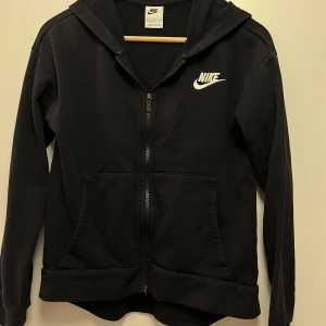 Svart hoodie från Nike - Säljer en svart hoodie från Nike i mycket bra skick. Den har en dragkedja framtill och en mysig fleecefodrad insida. Perfekt för höst och vinter! Den har långa ärmar och en klassisk Nike-logga på bröstet. Superbekväm och stilren för vardagsbruk. STOLEK 156 - 166