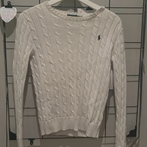 Vit stickad tröja från Ralph Lauren - Säljer en superfin vit stickad tröja från Ralph Lauren. Den har ett klassiskt flätmönster och den ikoniska loggan broderad på bröstet. Perfekt för både höst och vinter, och den är i mycket bra skick. Passar perfekt till jeans eller kjol för en stilren look! Orginalpris runt 1000kr