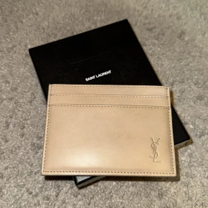Saint Laurent plånbok - Säljer ett elegant och stilrent kortfodral från Saint Laurent i en snygg beige färg. Perfekt för att hålla dina kort organiserade. Kortfodralet är i skinn och har en diskret YSL-logga i hörnet. Kommer med originalförpackning och dustbag. Perfekt för både vardag och fest!