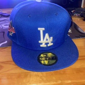 Blå keps från New Era - Säljer en blå New Era 59FIFTY keps med LA-logga framtill och blommigt broderi på baksidan. Kepsen är i bra skick och har en platt skärm. Perfekt för sommarens alla äventyr eller som en cool accessoar till vardagsoutfiten.