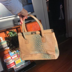 Beige och grön handväska i krokodilmönster - Säljer en superfin handväska i beige och grönt med krokodilmönster. Väskan har en klassisk form med två handtag och en snygg låsdetalj på toppen. Perfekt för både vardag och fest! Obs! Väskan har stått oanvänd länge och skinnet är därför ”kladdigt”💓